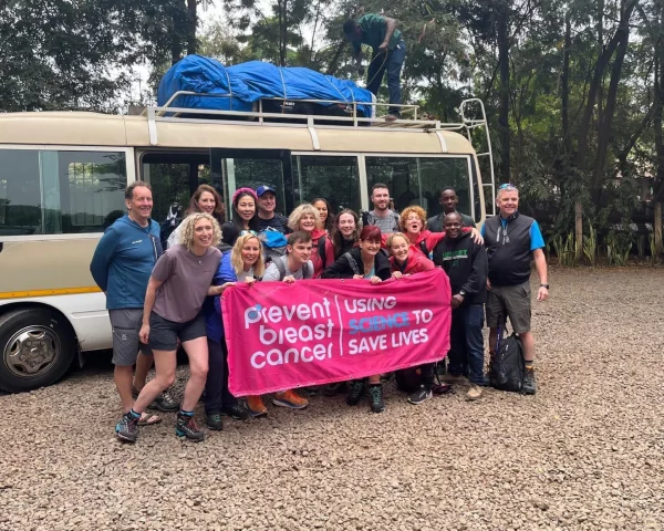 Group by minibus holding a pink banner that says 'Prevent breast cancer', with luggage strapped on the roof.