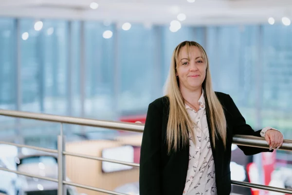 Woman with long blonde hair in a black blazer and patterned blouse leaning on a metal railing in a bright modern room.