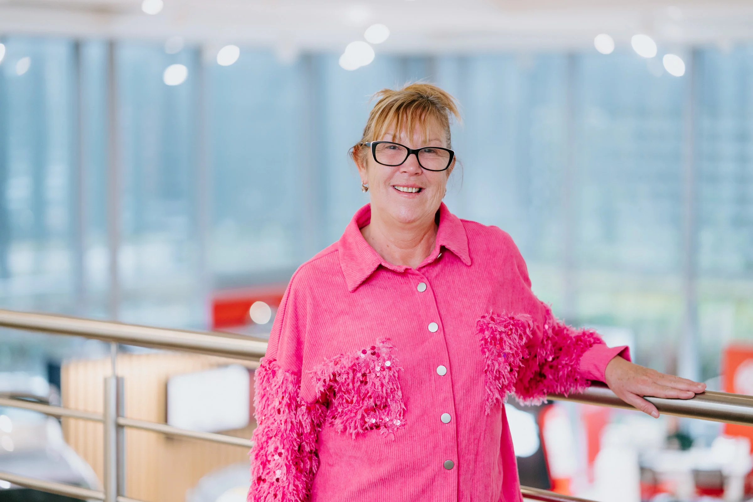Smiling woman wearing black glasses and a bright pink textured jacket, leaning on a metal railing in an indoor space.