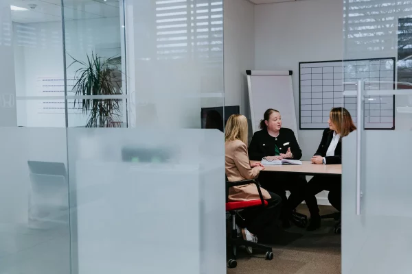 Three women in a glass-walled meeting room discuss documents at a table with a flipchart and whiteboard.