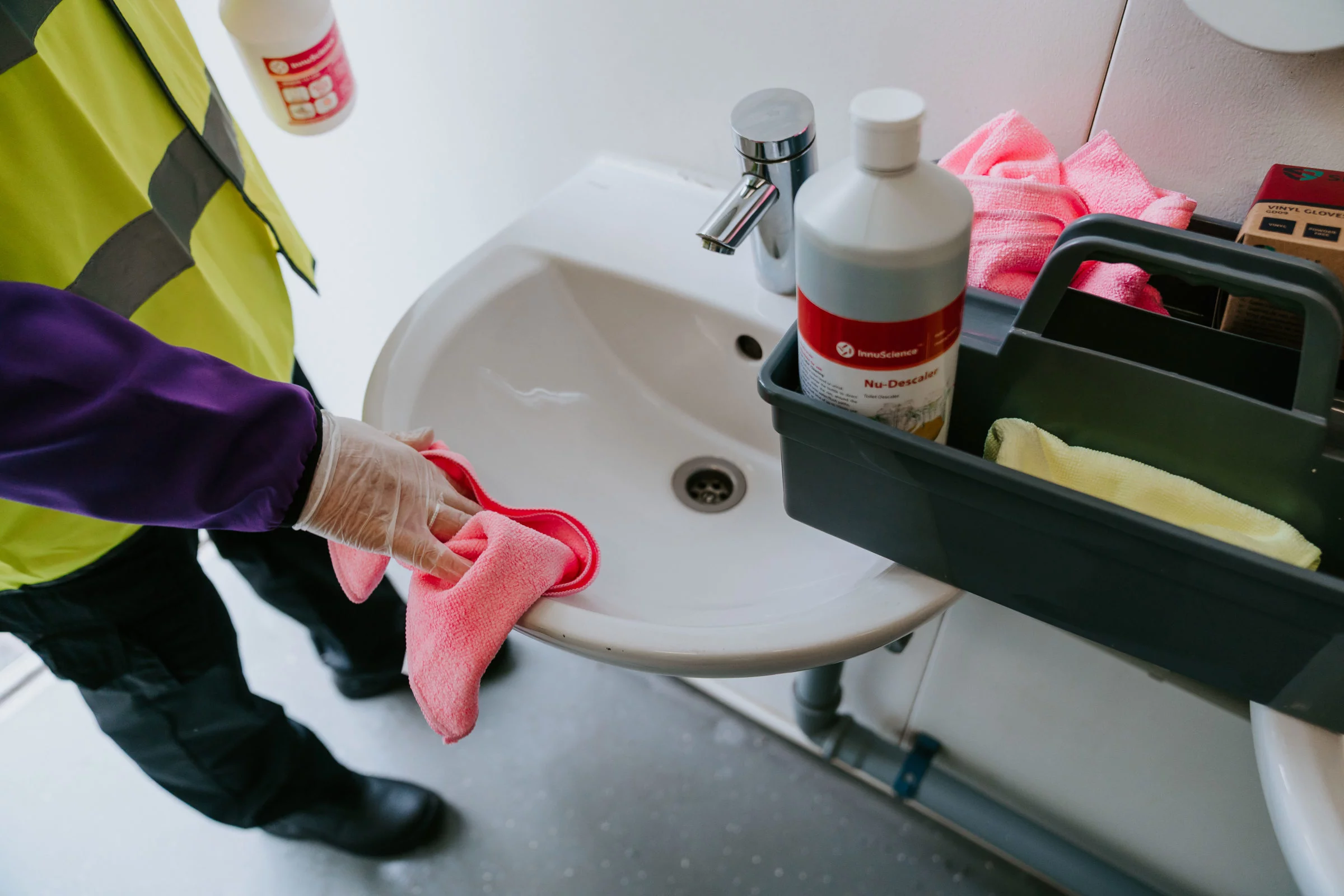 Gloved worker wiping a white sink with a pink cloth, plastic caddy with descaler bottle beside it.