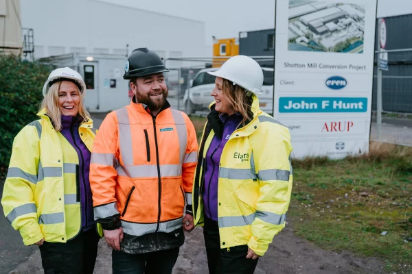Three construction workers in high-visibility jackets and hard hats smiling at a construction site with a project sign behind them.