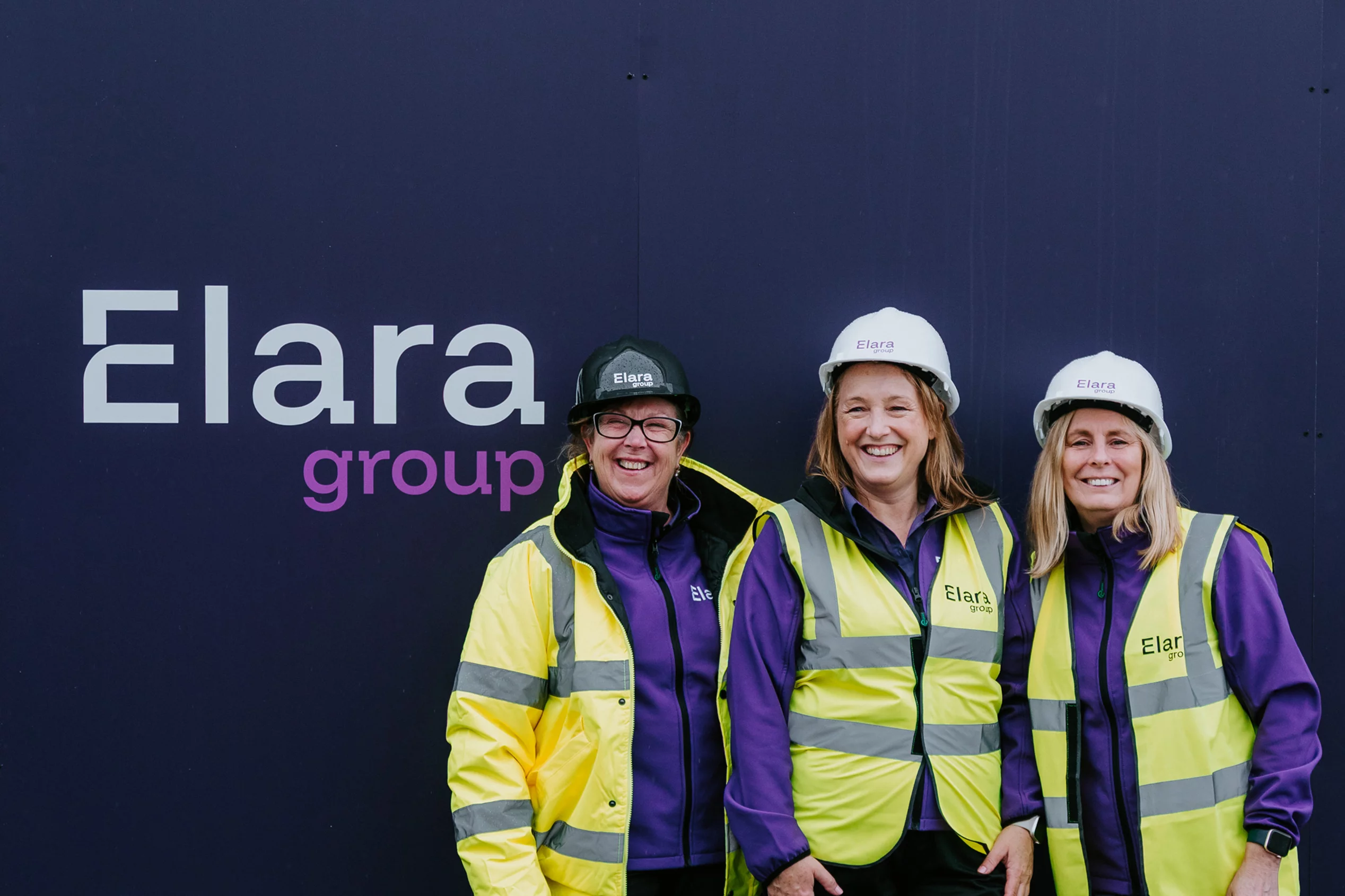Three women in hard hats and yellow safety vests smiling in front of a dark wall branded Elara group.