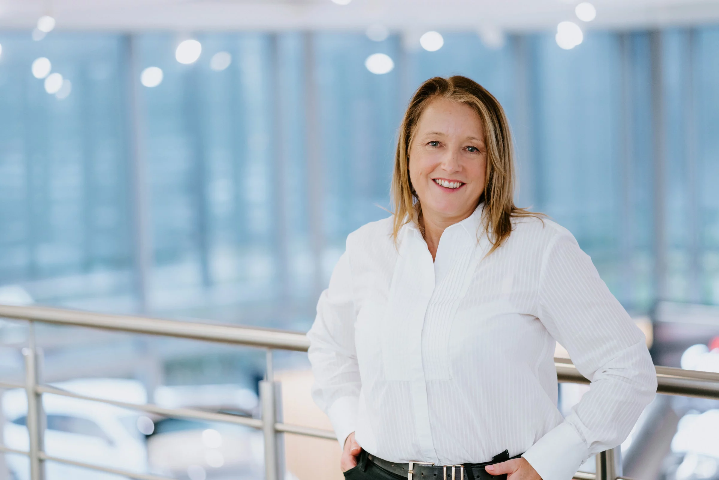 Smiling woman in a white shirt standing by a metal railing with hands on hips in a bright office atrium